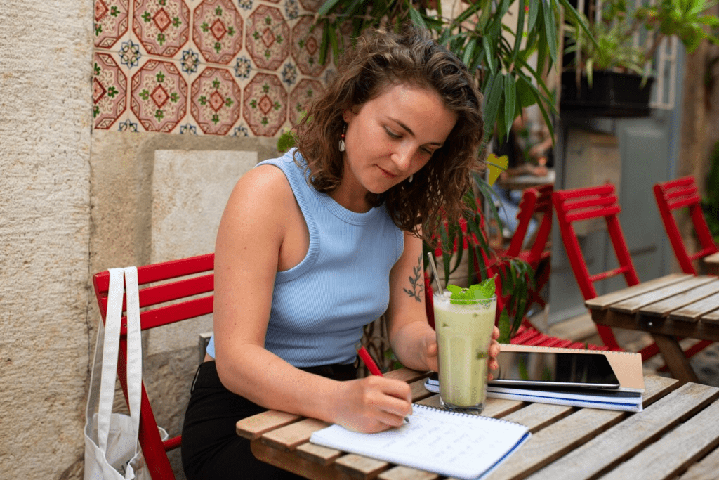 Mulher em cafeteria tomando suco e escrevendo no caderno.