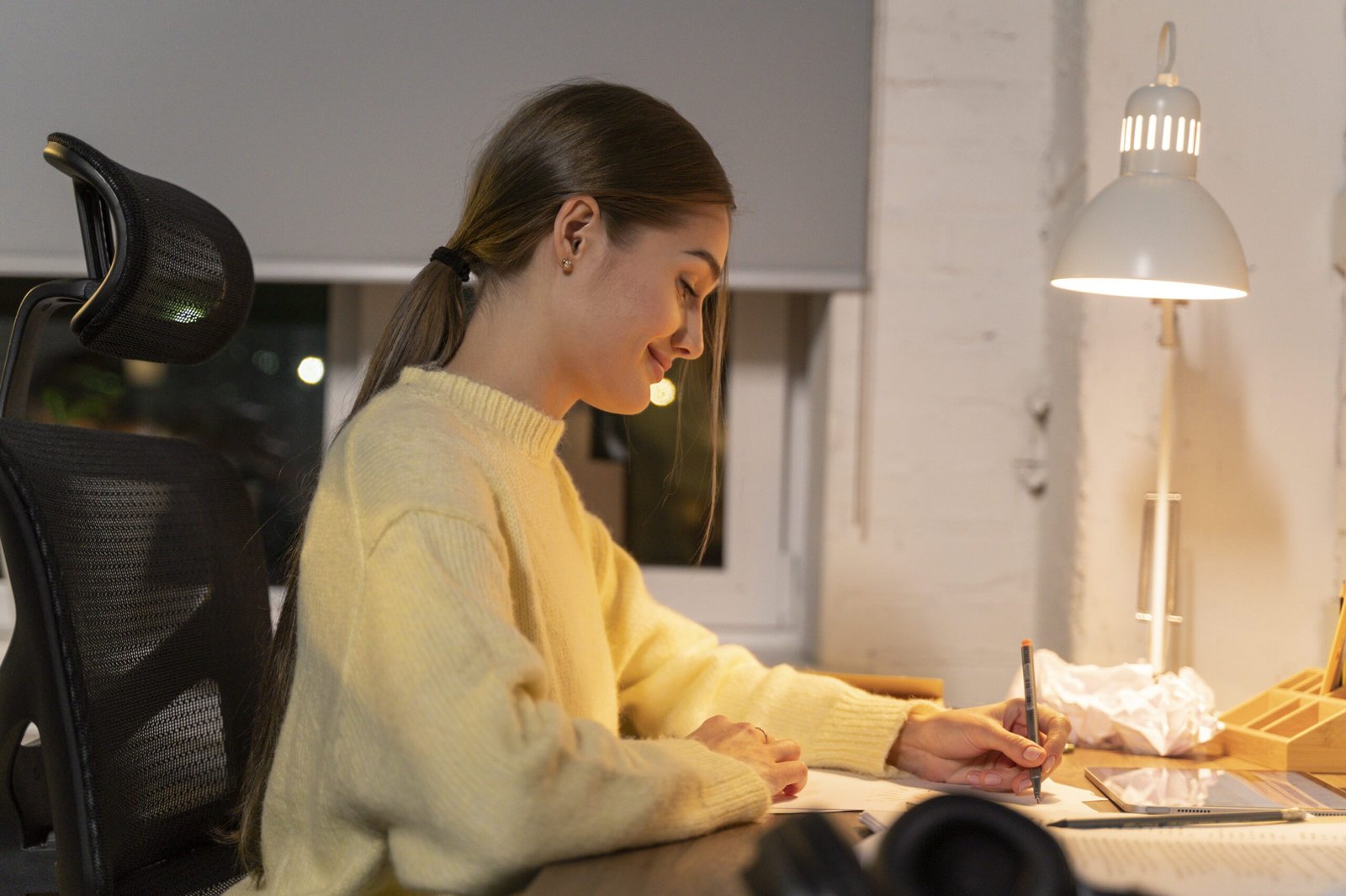 Mulher escrevendo a mão no papel, sentada em uma cadeira ergonômica, em seu escritório home office
