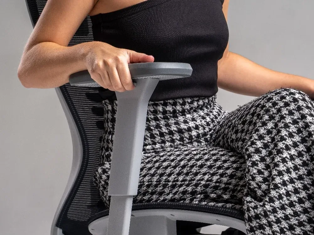 Garota jovem com blusa preta de manga única e calça xadrez sentada na cadeira ergonômica Elements Vertta Pro ajustando o apoio de braço - Melhor cadeira ergonômica da Elements