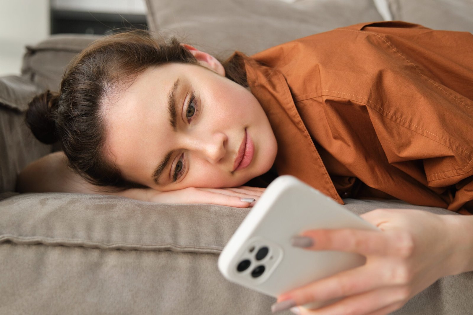 Mulher jovem deitada no sofá com a cabeça apoiada sobre uma das mãos, segurando o celular e vendo algum conteúdo - Slow content
