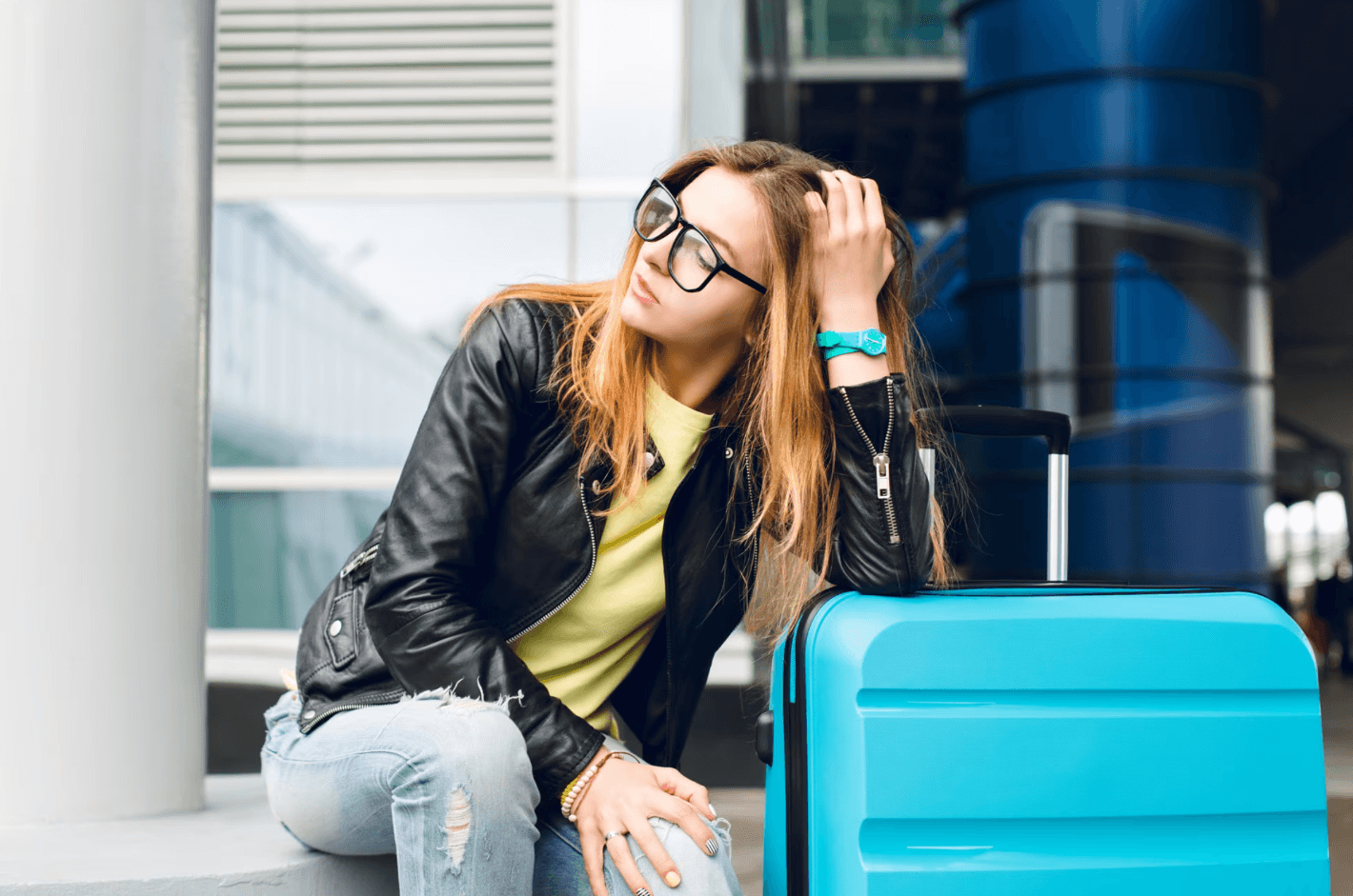 Mulher jovem e bonita de óculos escorada na mala azul, esperando viagem atrasada - Nômade digital