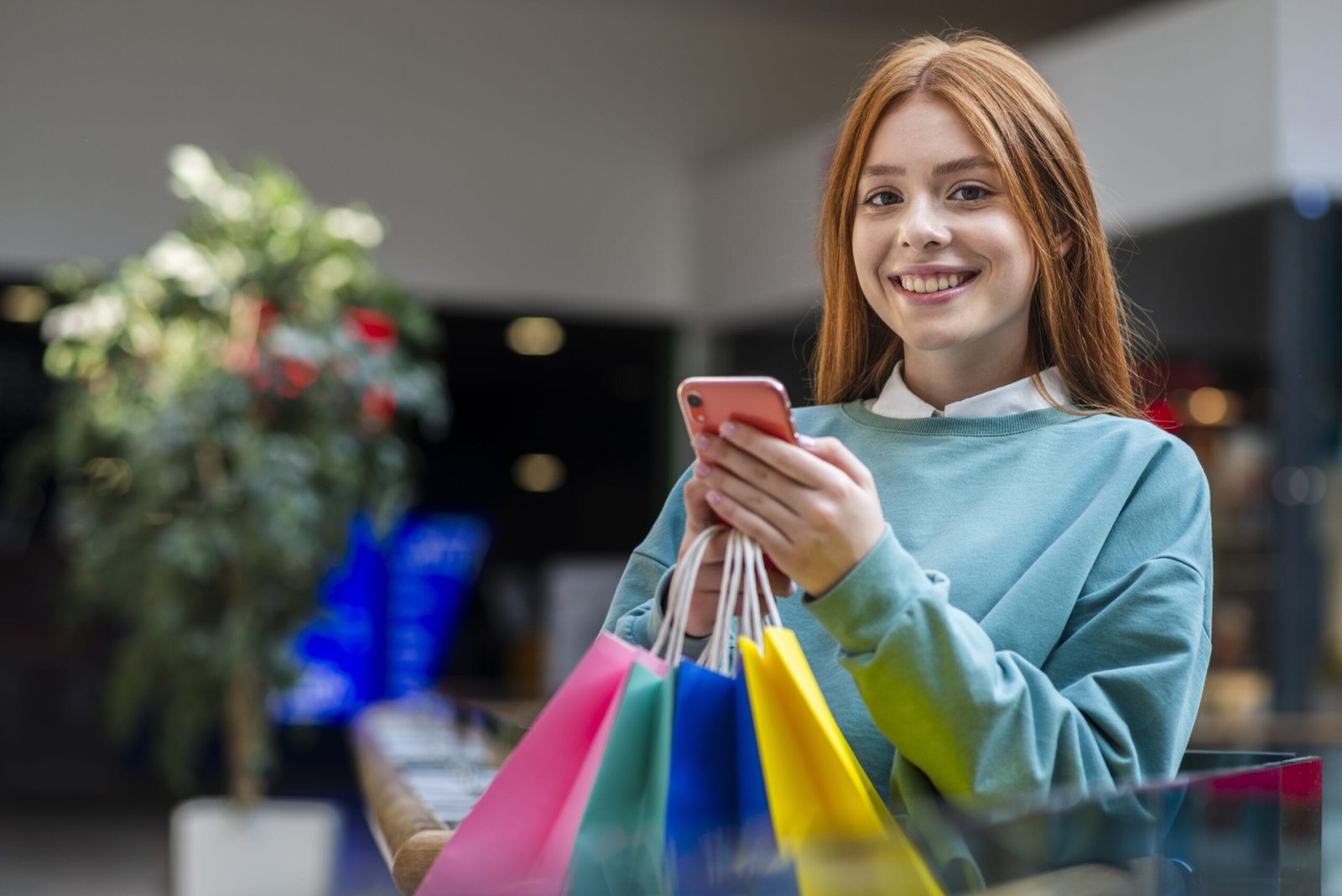 Garota ruiva bonita sorrindo usando o celular enquanto segura várias sacolas de compras - Comportamento do consumidor digital