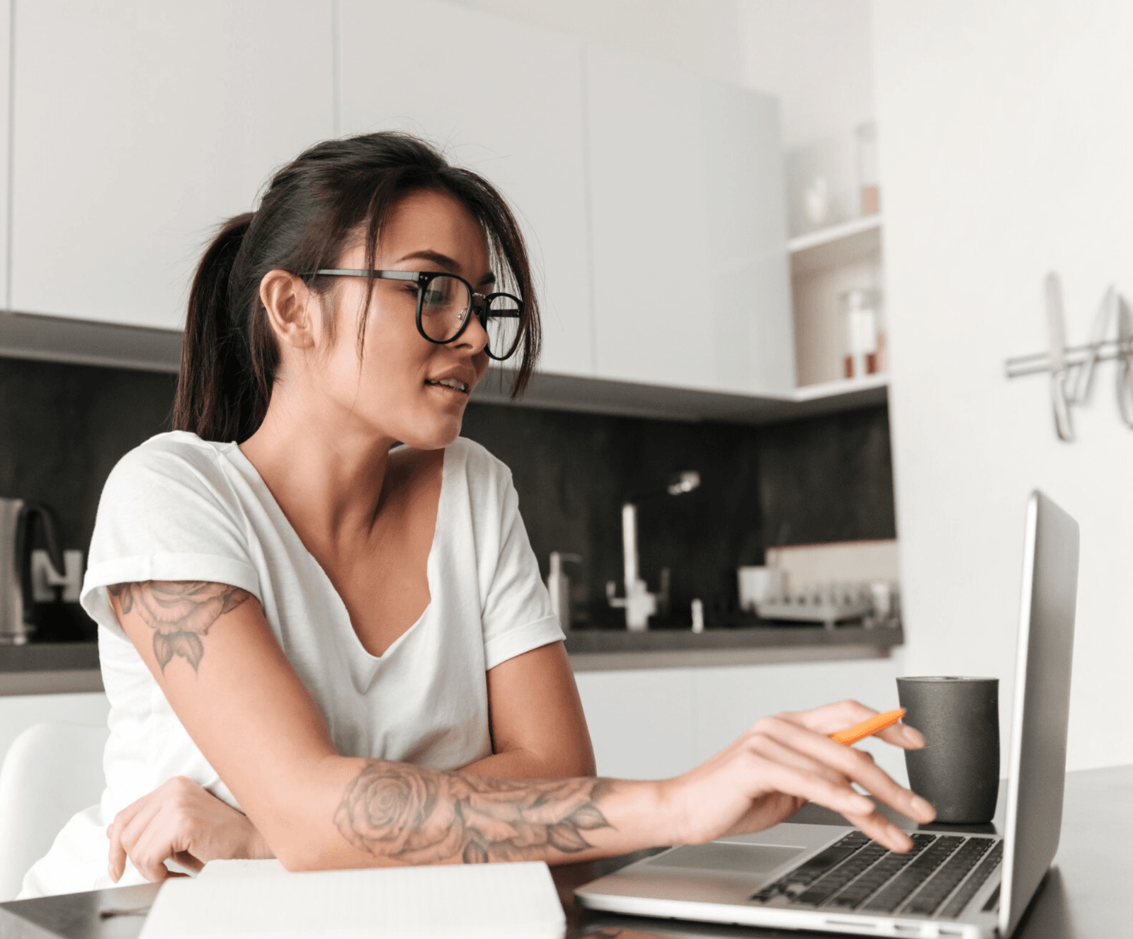 Mulher jovem e bela de óculos com tatuagens de flores no braço direito trabalha com o notebook em home office - Profissões home office mais bem pagas